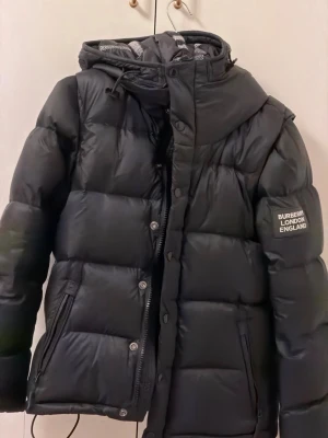 Svart pufferjacka från Burberry (jacket puffer to vest) - Snygg svart pufferjacka från Burberry med avtagbara ärmar så den kan bäras som väst. Jackan har huva med rutigt foder, dragkedja och knappar framtill samt logopatch på ärmen. Jackan blir också en väst. Obs! Sista bilden är inte min då ville bara visa hur de blir en väst! 