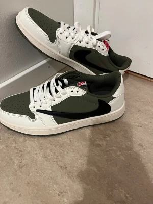 Nike Air Jordan 1 Low Travis Scott - Nike Air Jordan 1 Low Travis Scott i olivgrönt, vitt och svart med röd broderad logga och Travis Scott smiley på hälen. Skorna har snörning, platt sula och rund tå. Materialet är skinn och syntet, med ikonisk omvänd swoosh på sidan.