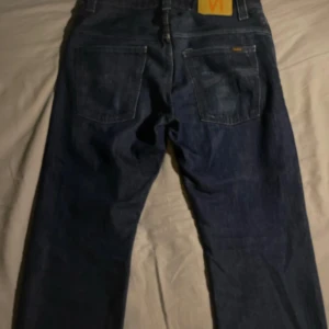 Mörkblå jeans från Nudie - Säljer ett par mörkblå jeans från Nudie med klassisk femficksdesign och raka ben. Jeansen har en snygg, enkel look med Nudie-loggan på bakfickan och är tillverkade i slitstarkt denim. Perfekt för dig som gillar stilrena och tidlösa jeans.