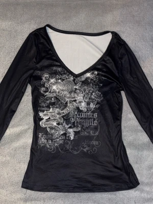 Svart långärmad topp med tryck - Säljer en svart långärmad topp med djup v-ringning och silverfärgat tryck framtill. Trycket har detaljer som hjärta, rosor och text, samt glittriga stenar som ger extra edge. Materialet är stretchigt och följsamt, perfekt för dig som gillar statement-plagg.