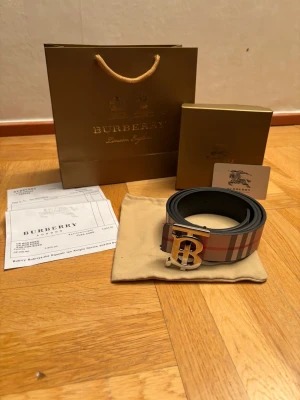 Rutig Burberry skärp med B-spänne - Snyggt Burberry skärp med klassiskt rutig design i beige, svart, vit och röd. Skärpet har ett stort metallspänne med Burberrys ikoniska B-logga och är tillverkat i äkta skinn. Perfekt för att lyfta din outfit med en lyxig touch.