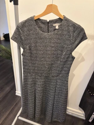 Gråmelerad kort klänning från H&M - Kort klänning från H&M i gråmelerat mönster med rund halsringning och korta ärmar. Klänningen har en figurnära överdel och lätt utställd kjol, samt dragkedja i ryggen. Perfekt för dig som gillar en stilren och enkel look.