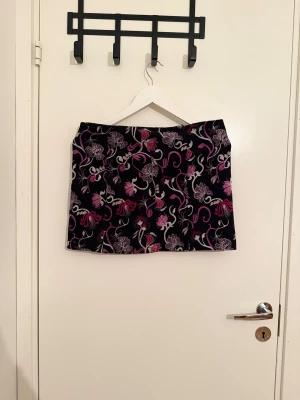 Blommig manchesterkjol från Zoul - Supersöt kort manchesterkjol från Zoul med blommigt mönster i lila, rosa och vitt på svart botten. Kjolen har en smidig dragkedja i sidan och är perfekt för dig som gillar färg och mönster. Snygg passform och skön känsla!