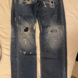 Dsquared2 blå slitna jeans, strl 42 - Säljer ett par blå jeans från Dsquared2 i storlek 42. Jeansen har flera stora och små hål, slitningar och en tvättad look som ger dem en riktigt edgy vibe. Klassisk femficksmodell med raka ben och grova detaljer. Perfekt för dig som gillar streetstyle.