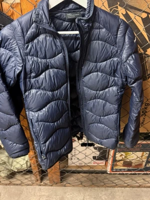 Mörkblå pufferjacka från Peak Performance - Säljer en mörkblå pufferjacka från Peak Performance med vågigt quiltad design och dragkedja framtill. Jackan har hög krage och långa ärmar, perfekt för kyliga dagar. Materialet är syntet och jackan har en clean, sportig look.