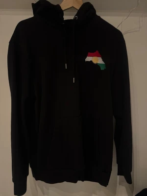 Kurdistan hoodie (broderad) - Svart hoodie med en broderad kurdisk flagga på bröstet. Hoodien har en justerbar dragsko vid huvan och en praktisk ficka på magen. Hoodien är iprincip ny, används max 2 gånger. Hoodien är liten i storleken, så denna känns som en M/L än XL.