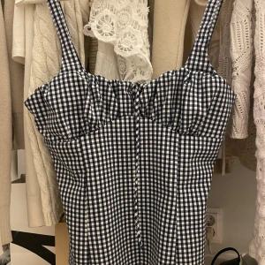 Rutig korsettopp med snörning - Supersöt korsettopp med smala axelband och rutig design i marinblått och vitt. Toppen har snörning framtill och markerad byst för en snygg siluett. Perfekt att styla med jeans eller kjol för en trendig look.