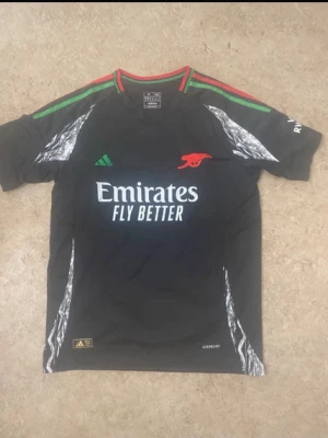 Arsenal Adidas svart matchtröja - Svart Arsenal matchtröja från Adidas med gröna och röda detaljer på axlarna, silvermönster längs sidorna och röd Arsenal-logga. Tröjan har trycket 'Emirates Fly Better' på bröstet och är tillverkad i lätt polyester för sportig känsla.