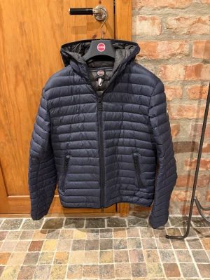 Colmar Jacka  - Snygg mörkblå pufferjacka från Colmar med huva och horisontella sömmar. Jackan har dragkedja framtill och två fickor med dragkedja. Perfekt för kyliga dagar och har en clean, sportig look.