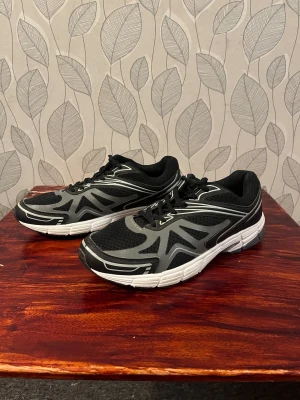 Svarta och grå sneakers med vit sula - Snygga svarta sneakers med grå detaljer och vit sula. Ovandelen är i mesh för bra andningsförmåga och skorna har klassisk snörning. Perfekta för dig som gillar sportig stil och vill ha bekväma skor till vardagen.