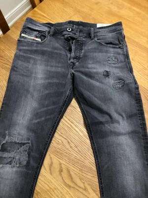 Svarta Diesel jeans med slitningar 32/33 - Säljer ett par svarta jeans från Diesel med coola slitningar och distressed detaljer på benen. Jeansen har klassisk femficksdesign, knappgylf och snygg Diesel Industry-märke vid fickan. Passformen är smal och materialet är mjukt jeans. Storlek 32/32 i mycket bra skick!