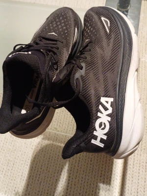 Svarta Hoka Clifton 9 sneakers - Säljer ett par Hoka Clifton 9 sneakers i svart med vita detaljer och tjock, vit sula. Skorna har snörning och ett mönstrat mesh-tyg som andas. Perfekta för dig som gillar att springa eller vill ha riktigt sköna sneakers med platt sula och rund tå.