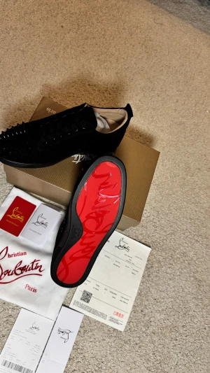 Christian Louboutin svarta sneakers med nitar - Svarta sneakers från Christian Louboutin med ikonisk röd sula och snörning. Tåpartiet är täckt av svarta nitar för en edgy look. Skorna är tillverkade i mocka och skinn, med rund tå och platt sula. Perfekt för dig som vill sticka ut med exklusiv design.