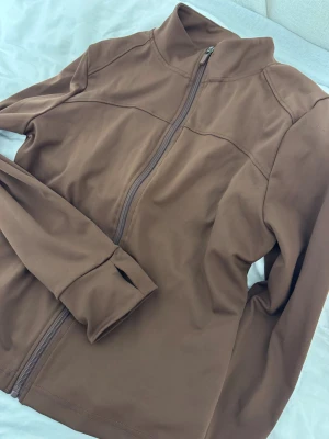 Brun zipjacka med lång ärm - Säljer en brun jacka med hel dragkedja och hög krage. Jackan har lång ärm med tumhål och är tillverkad i ett slätt, stretchigt material. Enkel och clean design som passar perfekt till lager-på-lager-stil.
