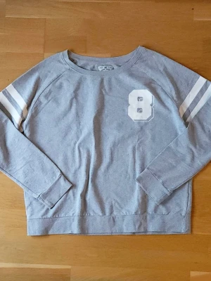 Grå sweatshirt med tryck - Grå sweatshirt med tryck på ärmarna och på bröstet. Tröjan har rund halsringning och långa ärmar. Perfekt för en avslappnad och sportig stil.