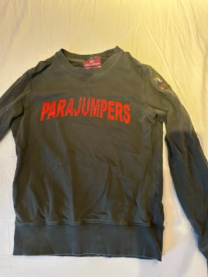  Parajumpers M - Snygg grå fleece-tröja från Parajumpers i storlek M. Stor röd logga på bröstet och patch på ena ärmen. Tröjan har rund hals och långa ärmar med ribbade muddar. Perfekt för dig som gillar streetwear och vill ha något bekvämt och stilrent.