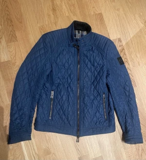 Blå quiltad jacka från Belstaff stl 48 män - Snygg mörkblå quiltad jacka från Belstaff med ståkrage och spänne, två dragkedjefickor fram och rutigt foder. Jackan har raka linjer, lång ärm och coolt emblem på armen. Perfekt för dig som gillar stilren design med sportig touch.