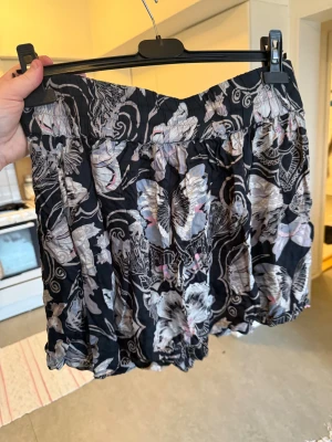 Svart blommig kortkjol med volang - Supersöt svart kortkjol med blommigt mönster i grått, vitt och ljusrosa. Kjolen har en luftig och volangig form med resår i midjan för skön passform. Perfekt för dig som gillar en lekfull och trendig stil. Från märket Odd Molly. Storlek 2 som enligt deras storleksguide motsvarar en M. Fin men det finns tyvärr en tråddraning. 