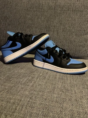Nike Air Jordan 1 Low blå/svart - Nike Air Jordan 1 Low i en snygg blå och svart färgkombination med vit mellansula. Skorna har klassisk snörning, rund tå och är tillverkade i skinn. Perfekt för dig som gillar streetstyle och vill sticka ut med en ikonisk sneaker. Med box