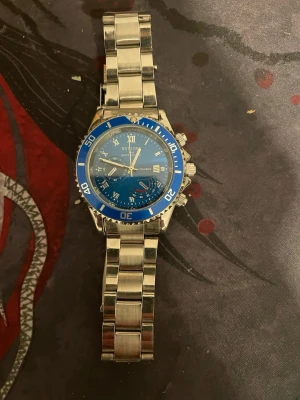 Blå och silverfärgad klocka från Notion - Snygg klocka från Notion med blå urtavla och silverfärgat armband i metall. Klockan har flera små visare, datumfunktion och en sportig bezel i blått med vita siffror. Perfekt accessoar för dig som gillar stilrena detaljer och metallarmband.