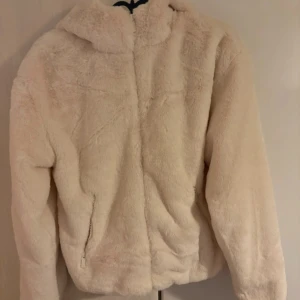 Beige teddyjacka med huva - Mysig teddyjacka i beige med fluffigt material och huva. Jackan har dragkedja framtill och två sidofickor med dragkedja. Perfekt för kalla dagar och har en loose passform som ger en chill vibe.