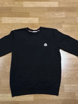 Svart sweatshirt från Moncler - Svart sweatshirt från Moncler med rund hals och diskret logotyp-patch på bröstet. Tröjan har långa ärmar och ribbade muddar vid hals, ärmslut och nederkant. Materialet är mjuk bomull som känns skön mot huden. Perfekt för dig som gillar stilrena och klassiska plagg.