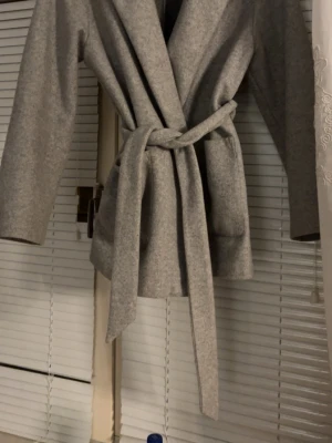 Grå omlott kappa med bälte Zara  - Snygg grå kappa med omlott-design och bälte i midjan. Jackan har breda slag, två stora fickor framtill och är långärmad. Perfekt för dig som gillar stilrena och enkla plagg med modern touch. Materialet känns mjukt och har en ull-liknande look.