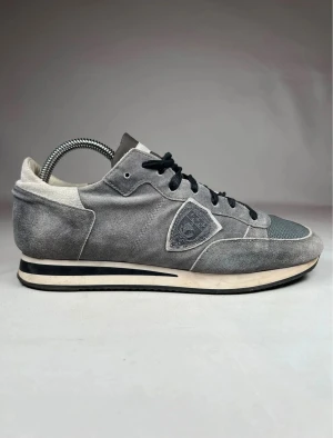 Grå Philippe Model sneakers mocka - Snygga grå sneakers från Philippe Model med svart snörning och rund tå. Ovandelen är i mjuk mocka med meshdetaljer framtill och en vit och svart randig sula. Märkets logga syns på sidan. Perfekt för dig som gillar stilrena och sportiga skor.