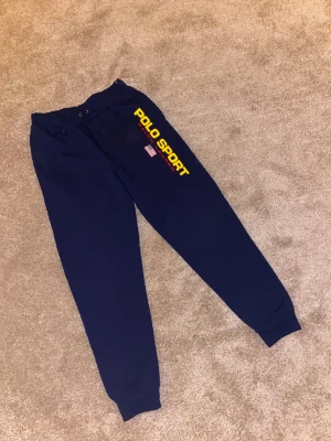 Mörkblå Polo Sport mjukisbyxor - Mörkblå mjukisbyxor från Polo Sport Ralph Lauren med gul och röd logga längs benet och amerikansk flagga. Byxorna har snörning i midjan och muddar vid bensluten. Perfekta för chill dagar eller när du vill ha en sportig look.