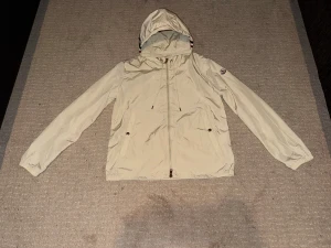 Beige vindjacka från Moncler med huva - Snygg beige vindjacka från Moncler med huva och dragsko. Jackan har hel dragkedja framtill, två fickor med tryckknappar och Moncler-logga på ärmen. Huvan har coola röd, vit och marinblå ränder. Perfekt för dig som vill ha en stilren och sportig look.