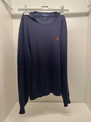 Marinblå tröja från Polo Ralph Lauren - Snygg marinblå tröja från Polo Ralph Lauren i slim fit med orange broderad logga på bröstet. Tillverkad av mjuk pima bomull, rund hals och långa ärmar. Perfekt för dig som gillar klassisk stil med en modern touch.