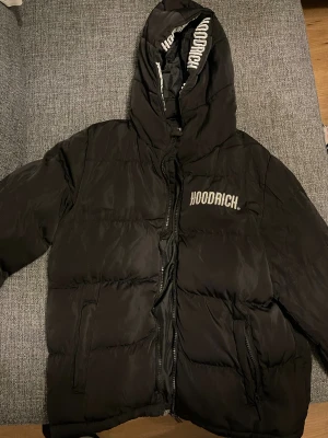Svart pufferjacka från Hoodrich - Svart pufferjacka från Hoodrich med stor huva och broderad logga på bröstet och huvan. Jackan har dragkedja framtill och två sidofickor. Perfekt för kalla dagar och har en cool streetstil med tydliga Hoodrich-detaljer.