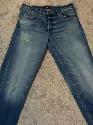Blå Lee jeans med slitningar - Säljer ett par klassiska blå jeans från Lee i storlek 30x32, med snygga slitningar på låret och knät. Jeansen har fem fickor, normal passform och raka ben. Materialet är robust denim i bomull och färgen är ljusblå med tvättade detaljer. Perfekta för en avslappnad och trendig look.