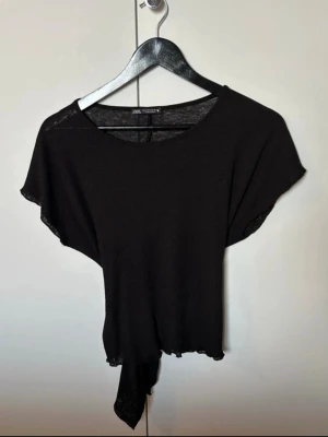 Svart knytblus från Zara, S - Svart blus från Zara i storlek S med korta ärmar och rund hals. Toppen har en snygg knytning nedtill i fram och bak, vilket ger en cool och avslappnad vibe. Materialet är tunt och mjukt, perfekt för varmare dagar. Enkel och stilren design som funkar till många looks.