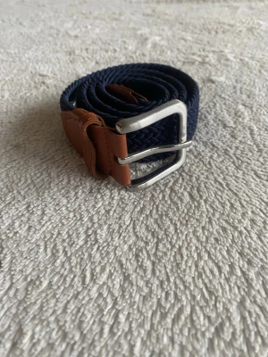 Marinblå flätad bälte från Jack & Jones - Säljer ett marinblått flätat bälte från Jack & Jones med snygga bruna skinn-detaljer och silverfärgat spänne. Perfekt accessoar för att lyfta din outfit med en clean och stilren look. Passar midjemått 90-92 cm.