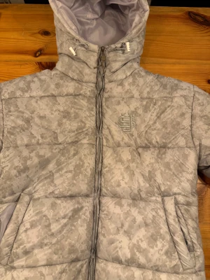 Vit Suevene pufferjacka  - Säljer den virala suevene. pufferjacakan .med de vita flagemönster. Jackan har huva med snörning, dragkedja framtill och två sidofickor. Fodret är ljusgrått och jackan är tillverkad i syntetmaterial. Perfekt för kalla dagar efter som den håller värmen och riktigt snygg streetstil. Storlek small men jag är 178 och sitter perfekt Använd typ 5 gånger 