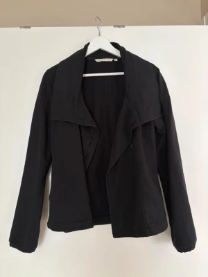 Svart kappa kort - Svart kavajjacka från House of Lola i medium med snygg bred krage och asymmetrisk knäppning framtill. Jackan har lång ärm och är tillverkad i mjukt bomullsmaterial. Perfekt för dig som gillar stilrena och enkla plagg med en twist.