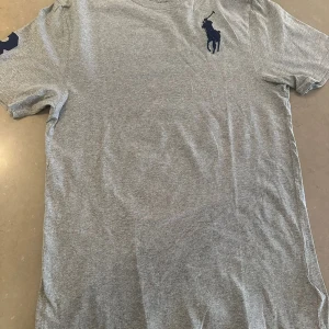 Grå t-shirt från Polo Ralph Lauren XL - Snygg grå t-shirt från Polo Ralph Lauren i storlek XL. Klassisk modell med korta ärmar och stort mörkblått broderat logotyp på bröstet. Tillverkad i mjuk bomull som känns skön mot huden. Perfekt för en clean och stilren look.