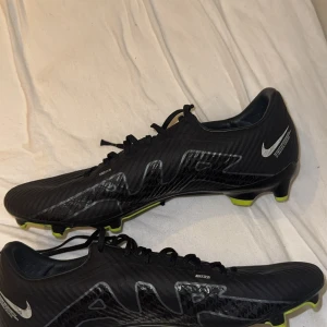 Nike Mercurial fotbollsskor svart/vit - Säljer ett par Nike Mercurial fotbollsskor i svart med silverdetaljer och gröna dobbar. Skorna har snörning, och en smidig, sportig design. Perfekta för dig som vill ha grymt grepp och lätt känsla på planen. Och den är använd max 4 gånger. 