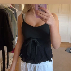 Svart linnetopp med knyt och volang - Snygg svart linnetopp med smala axelband och dekorativ knytning framtill. Toppen har volangkant nedtill och lätt rynkad effekt för en feminin look. Perfekt till jeans eller kjol för en trendig stil.