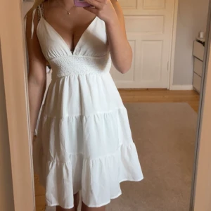 Vit kort sommarklänning med knytband - Supersöt vit kort klänning med smock i midjan och volangig kjol. Djup v-ringning och tunna axelband som knyts upptill. Perfekt för varma dagar och ger en fräsch och luftig känsla. Materialet känns lätt och passar perfekt till sommaren. Använd 1 gång