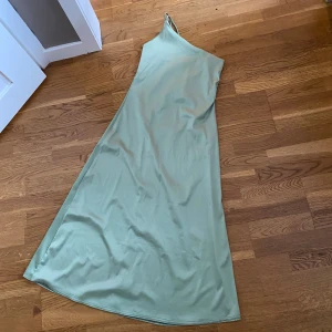 Ljusgrön långklänning med oneshoulder - Snygg ljusgrön långklänning med oneshoulder och tunna axelband. Klänningen har en enkel och stilren design med lätt utställd nederdel. Perfekt för dig som gillar minimalistisk stil och vill sticka ut med en cool asymmetrisk look.