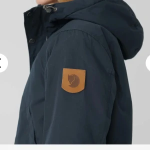 Svart parkajacka från Fjällräven - Svart parkajacka från Fjällräven med klassisk huva, stora fickor och snygga silverfärgade knappar. Jackan har det ikoniska läderloggan på ärmen och är tillverkad i slitstark bomull och polyesterblandning. Perfekt för kalla dagar och outdoor vibes.