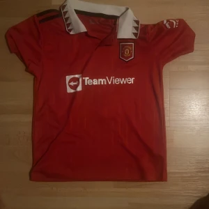 Manchester United Ronaldo 7 matchtröja - Röd Manchester United fotbollströja med Ronaldo 7-tryck på ryggen, TeamViewer och DXC Technology-loggor. Klassisk vit krage med svarta detaljer och klubbmärke på bröstet. Kortärmad modell i lätt och ventilerande material, perfekt för fotbollsälskare.