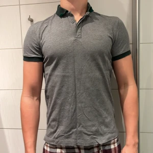 Hugo boss piké  - Snygg grå pikétröja från BOSS med mörkgrön krage och ärmslut. Klassisk design med knappar vid halsen och diskret BOSS-logga på bröstet. Tillverkad i mjukt bomull. Tröjan är 10/10 skick och har används ett fåtal gånger