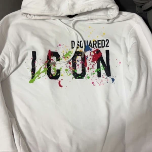 Vit hoodie Dsquared2 ICON färgstänk - Vit hoodie från Dsquared2 med stor svart 'ICON'-text och färgglada färgstänk i grönt, rött, gult, blått och rosa. Tröjan har huva med snörning och en avslappnad passform. Perfekt statement-plagg för dig som gillar streetstyle.