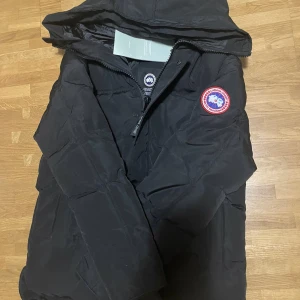 Svart dunjacka från Canada Goose M - Säljer en svart dunjacka från Canada Goose med huva och klassisk logga på ärmen. Jackan har dragkedja framtill och quiltad design som håller värmen. Perfekt för kalla dagar och riktigt snygg streetstil.