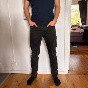  Nudie jeans  - Säljer ett par svarta jeans från Nudie med klassisk femficksdesign och raka ben. Jeansen har en mörk tvätt och subtila slitningar för en avslappnad look. Tillverkade i mjukt denimtyg med normal passform och medelhög midja.