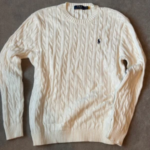 Vit kabelstickad tröja Polo Ralph Lauren - Vit kabelstickad tröja från Polo Ralph Lauren med klassisk rund hals och broderad marinblå logga på bröstet. Tröjan har ribbade muddar och är tillverkad i mjuk bomull. Perfekt för en clean och stilren look.