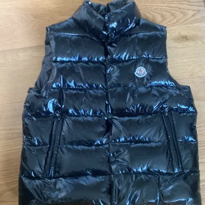 Svart dunväst från Moncler - Snygg svart dunväst från Moncler med glansig finish och hög krage. Västen har två praktiska fickor med dragkedja och ikonisk Moncler-logga på bröstet. Perfekt för lager-på-lager och streetstyle. Fodret har en cool serietidningsdetalj. Storlek 1 på västen och it hundra att den är riktig och därav priset.